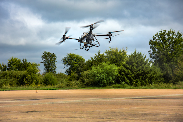 A 4 propeller drone hovers over pavement on a cloudy day in front of a backdrop of green trees and cloudy skies
