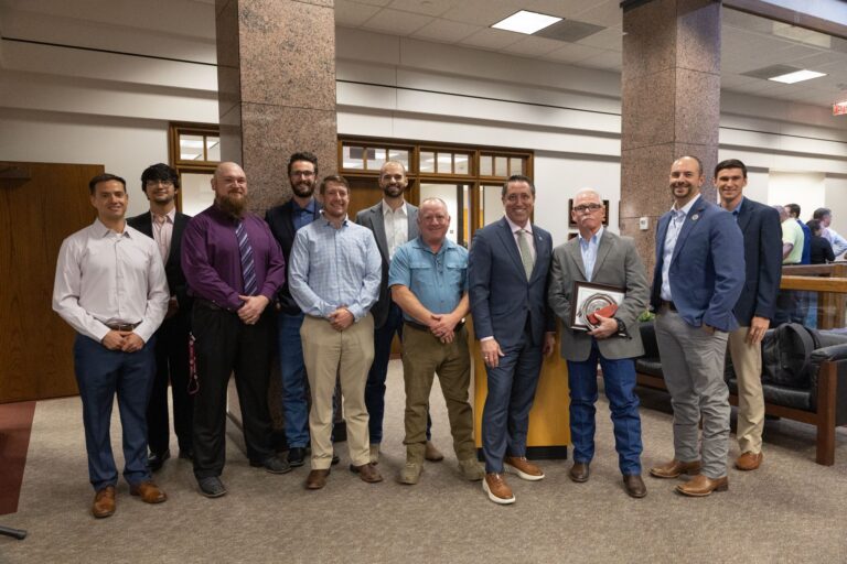 11 Men standing in a room, one man holding awards presented at an awards ceremony.