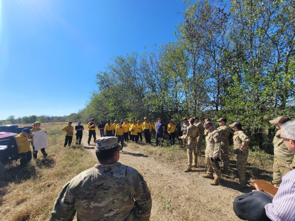 Roughly 30 people standing in a field, some are in military camouflage uniforms, others in firefighting gear.