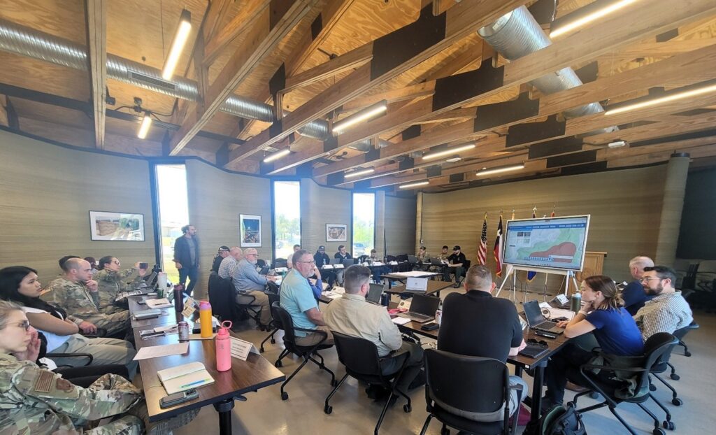 Approximately 20 people sitting in a conference room, a mix of military servicemembers and civilians looking at a screen at the front of the room.