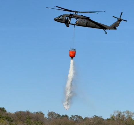 A UH-60 Blackhawk helicopter in flight with a water bucket attachment dropping water during a training exercise.