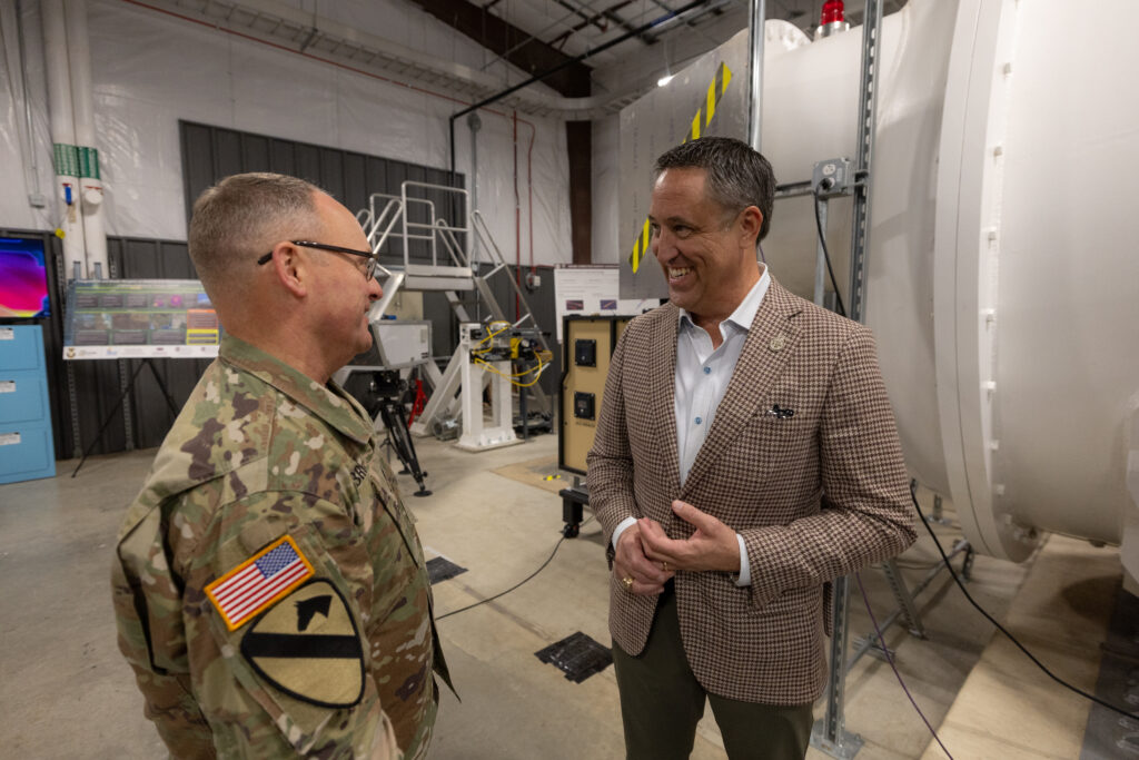 Two individuals, one in military uniform and the other in a suit, engage in conversation within a technical facility.